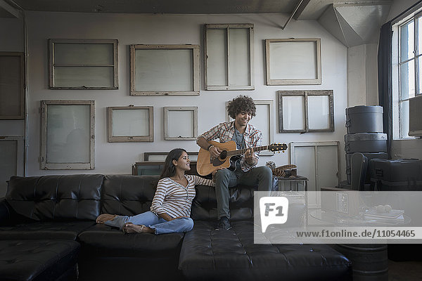 Loft-Dekor. Eine Wand  an der Bilder in Rahmen aufgehängt sind  die umgekehrt die Rückseiten zeigen. Ein Mann spielt Gitarre und eine Frau sitzt auf einem Sofa.