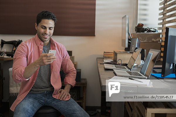 A man sitting in a home office  with two laptops on the desk  checking his smart phone.