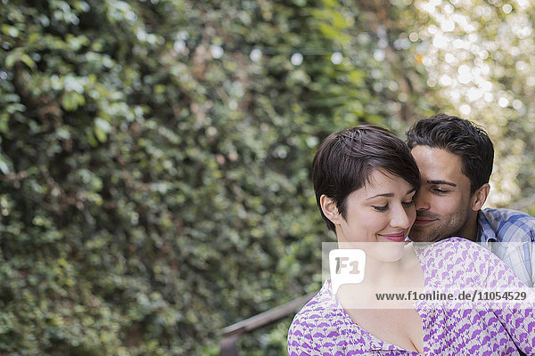 A couple in a city park beside a green wall of foliage embracing.