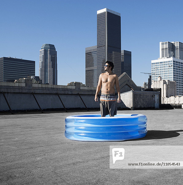A man standing in a small inflatable water pool on a city rooftop.