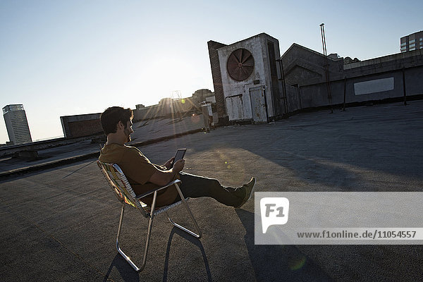 Ein Mann sitzt in einem Strandkorb auf dem Dach einer Stadt und benutzt ein digitales Tablet.