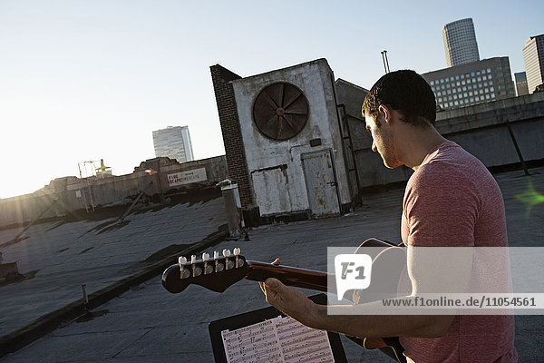 A man playing a guitar  sititng on a rooftop terrace overlooking the city at dusk.