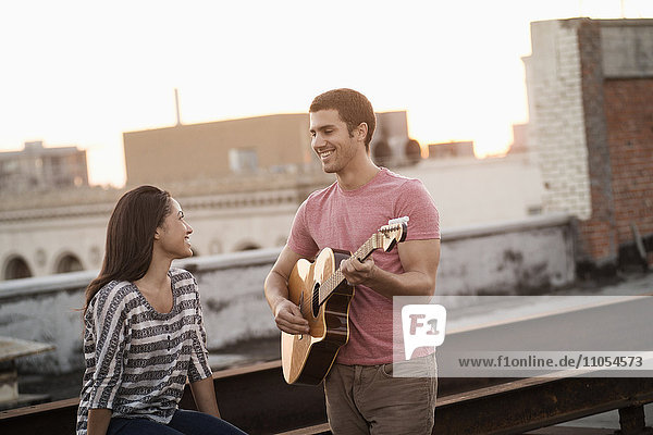 A man playing a guitar to a woman  on a rooftop terrace overlooking a city at dusk.