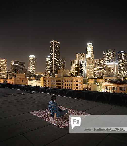 A man sitting on a rug on a rooftop overlooking a city lit up at night.