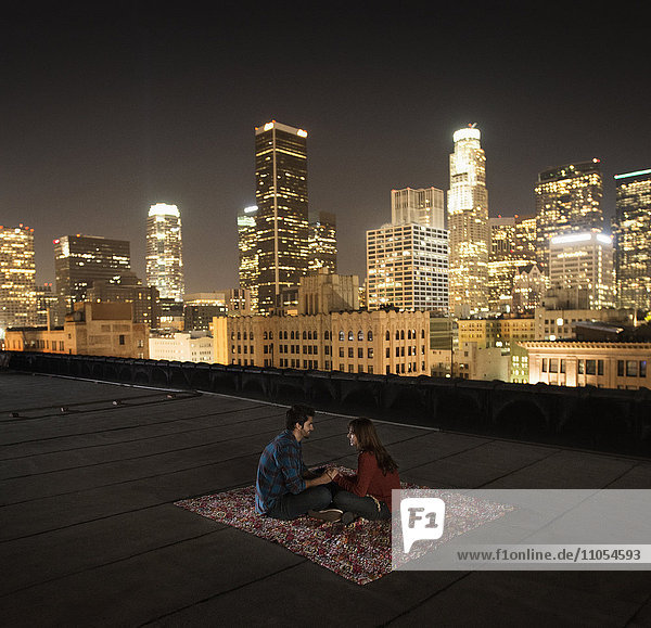 A couple sitting on a rug on a rooftop overlooking a city lit up at night.