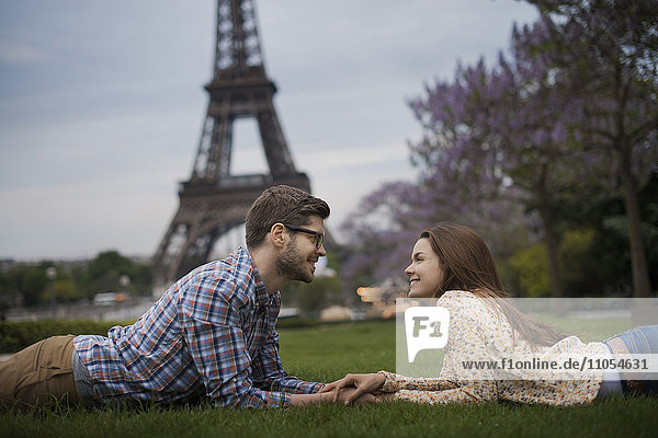 Ein Paar liegt auf dem Gras und hält sich im Schatten des Eiffelturms in Paris an den Händen.