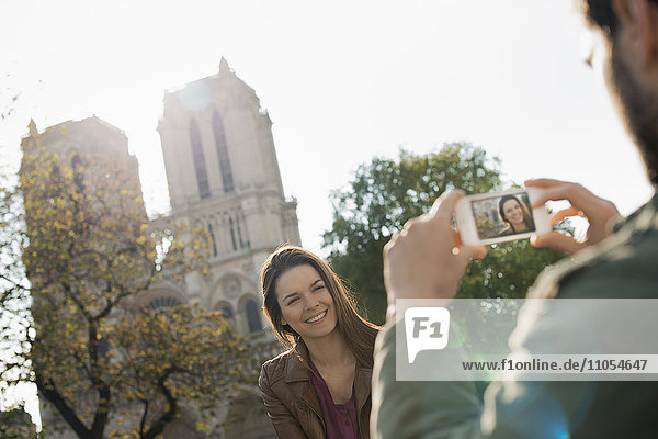 Ein Mann hält ein Smartphone in der Hand und fotografiert eine Frau vor der Kathedrale Notre Dame.