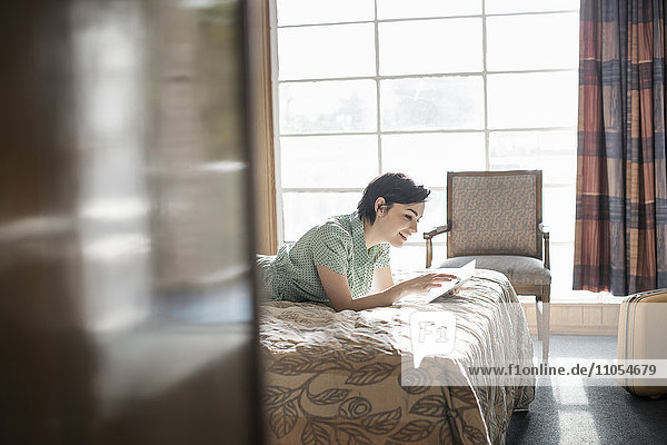 Eine junge Frau liegt auf einem Bett in einem Motelzimmer und benutzt einen digitalen Tablet-Touchscreen.