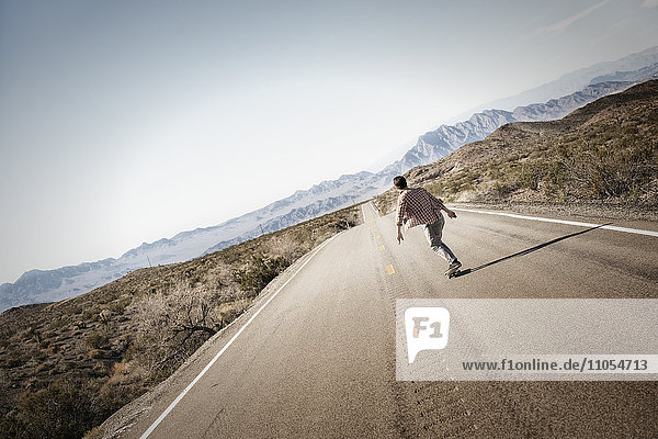 A young man riding down a tarmac road in the desert on a skateboard.