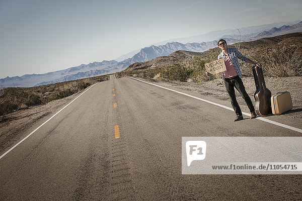 A man standing by the roadside  a hitchhiker with guitar and case  holding a sign saying Vegas or Bust.