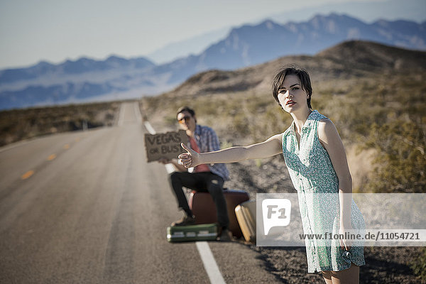 Ein junges Paar  Mann und Frau  auf einer asphaltierten Straße in der Wüste beim Trampen  mit einem Schild mit der Aufschrift Vegas oder Bust.