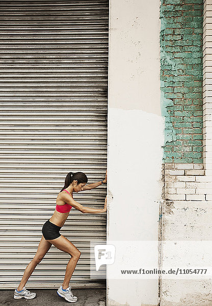 A woman in running gear  crop top and shorts  stretching her body and preparing for a run  or cooling down after exercise  by a steel shutter.