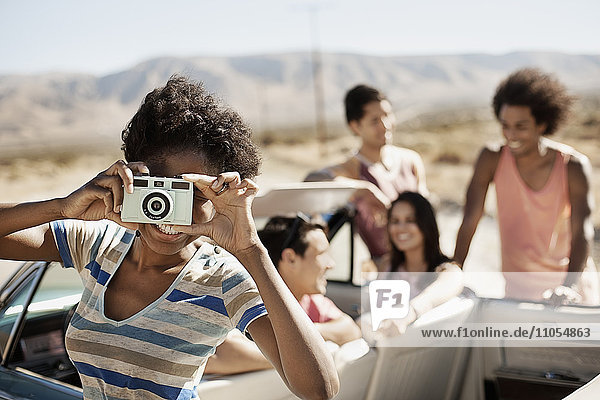 A group of friends by a pale blue convertible on the open road  on a flat plain surrounded by mountains  one holding a camera.