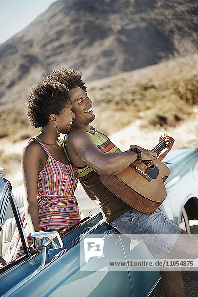 A young couple standing by a pale blue convertible on the open road  the man playing a guitar.