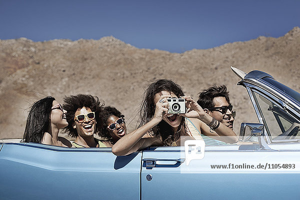 A group of friends in a pale blue convertible on the open road  driving across a dry flat plain surrounded by mountains.