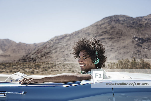A young man with music headphones sitting in the back of a pale blue convertible on the open road in the mountains.