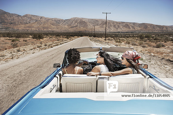 Three young people in a pale blue convertible car  driving on the open road across a flat dry plain
