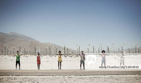 Eine Reihe von sechs jungen Leuten  die mit ausgestreckten Armen am Straßenrand vor der Kulisse von Windturbinen stehen.