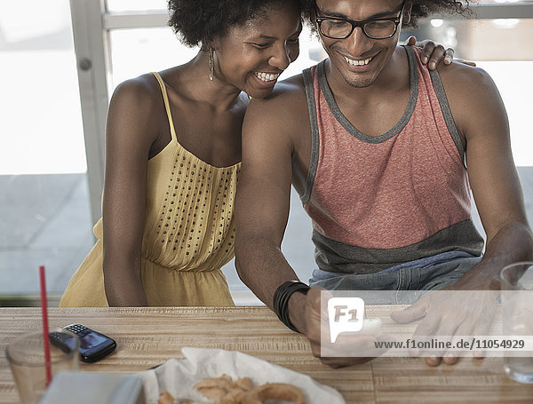 Ein junges Paar sitzt zusammen  umarmt sich und lacht über einen Bildschirmtext oder ein Bild auf einem Smartphone.