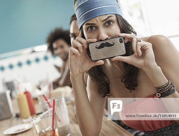 A woman holding a picture of a moustache on her smart phone just under her nose.