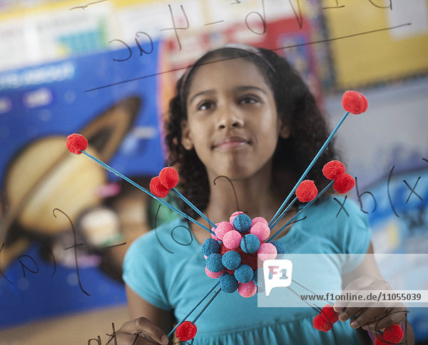 Ein Mädchen  das eine Molekularstruktur in der Hand hält und im Klassenzimmer auf eine Tafel mit Gleichungen und Formeln schaut.