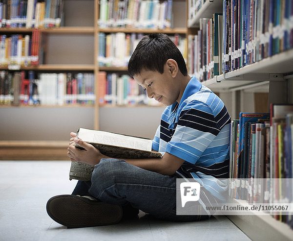 A boy sitting on the floor in a library reading a book.
