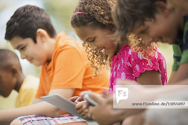 A row of children sitting outdoors in summer using tablets and handheld games.
