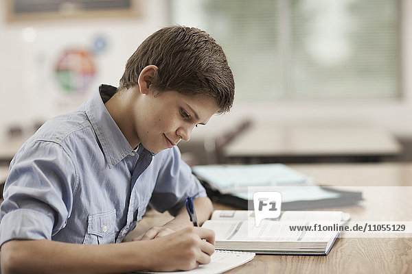 A boy sitting at a desk in class using school books and holding a pen.
