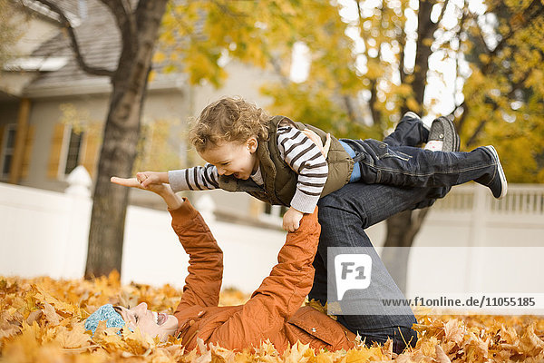 Eine auf Herbstlaub liegende Frau hält ein Kind in der Luft  das auf den Knien balanciert.