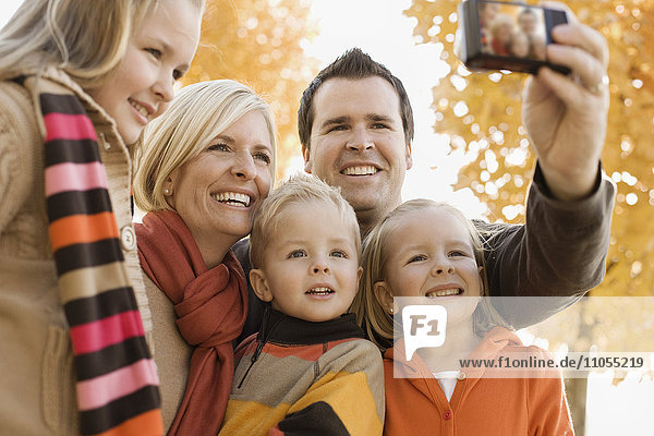 Eine Familie mit zwei Eltern und drei Kindern posiert für ein Selfy unter dem Herbstlaub an den Bäumen.