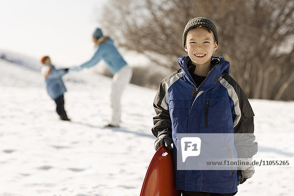 Ein Junge  der einen Schlitten im Schnee hält  und hinter ihm eine Mutter mit Kind.
