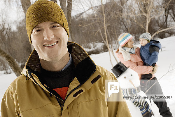 A man smiling in the foreground  and a woman holding a child beside a snowman.