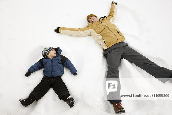 Two people  a man and a child lying in the snow make snow angel shapes.