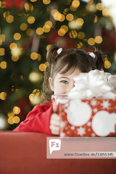 Ein Mädchen von einer Weihnachtspresse  das sein Gesicht hinter einem großen Paket versteckt.
