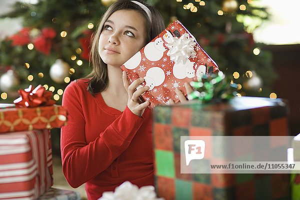 Ein Mädchen an einem Weihnachtsbaum schüttelt ein Geschenk und versucht zu erraten  um was es sich handelt.