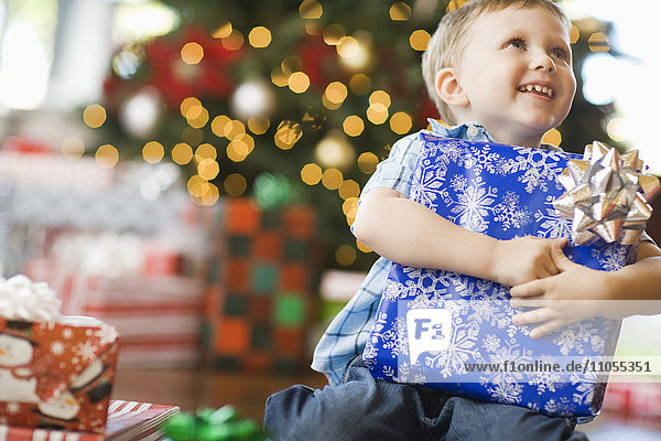 Ein kleiner Junge an einem Weihnachtsbaum  der ein großes eingepacktes Geschenk umarmt.