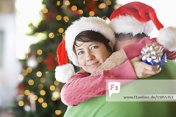 Ein Paar mit Nikolausmützen  das sich vor einem Weihnachtsbaum umarmt.