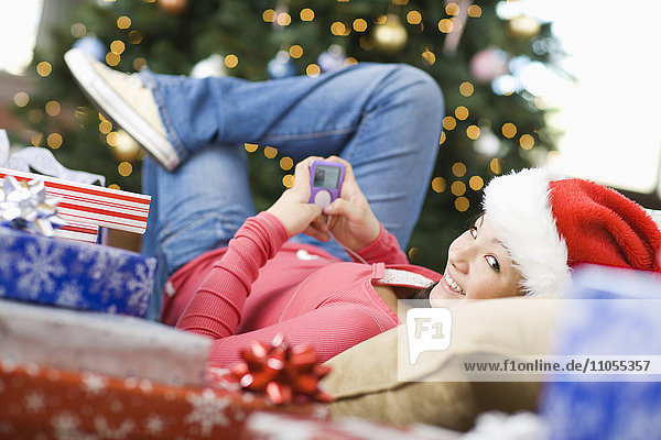Ein Mädchen mit Weihnachtsmannmütze  das mit einem Mobiltelefon telefoniert und vor einem Weihnachtsbaum liegt.