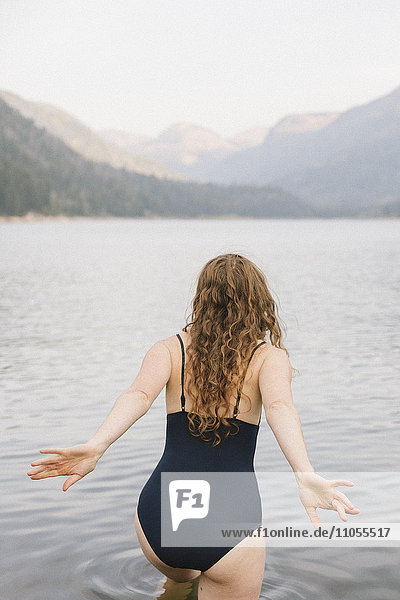 Eine Frau in einem schwarzen Badeanzug  die in einen ruhigen See in den Bergen watet.