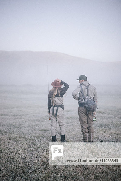 Two people walking across a meadow in early morning mist carrying fishing rods.