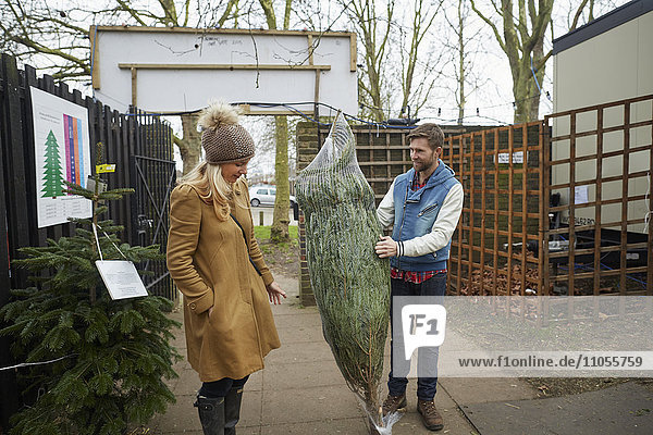 A member of staff carrying a traditional pine tree  Christmas tree talking to a woman client.