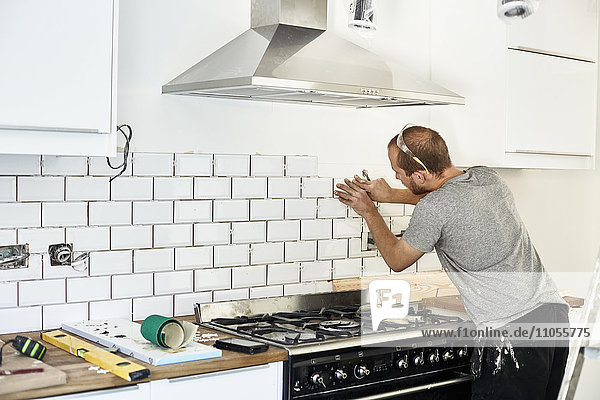 A man working in a new kitchen  a tiler applying tiles to the wall behind the cooker.