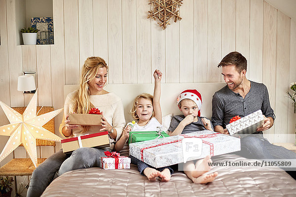 A family  two adults and two children sitting in bed on Christmas morning opening presents together.