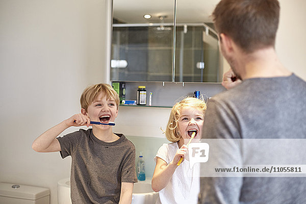 Two children and an adult man cleaning their teeth with toothbrushes in a bathroom.