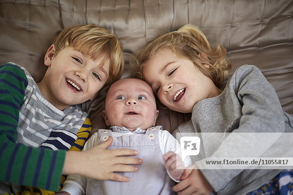 Drei Kinder liegen auf einem Bett  ein Junge und ein Mädchen mit einem Baby dazwischen.