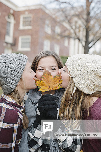 Drei junge Mädchen stehen in einer Reihe  eines hält ihr ein Herbstblatt vor das Gesicht  zwei küssen sie auf die Wange.