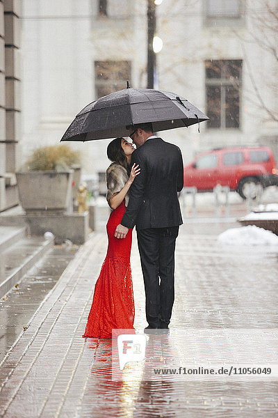 Eine Frau in einem langen roten Abendkleid mit Fischschwanzrock und Pelzstola und ein Mann im Anzug  der sich auf einer Straße unter einem Regenschirm küsst.