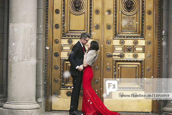 A woman in a long red evening dress with fishtail skirt and a fur stole  and a man in a suit kissing on the steps of a building.