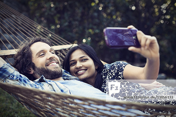 Ein Ehepaar  ein junger Mann und eine junge Frau  die in einer großen Hängematte im Garten liegen und sich ein Selfy von sich selbst nehmen.