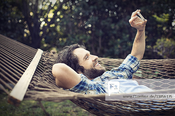 A man lying in a garden hammock taking selfies with his phone.
